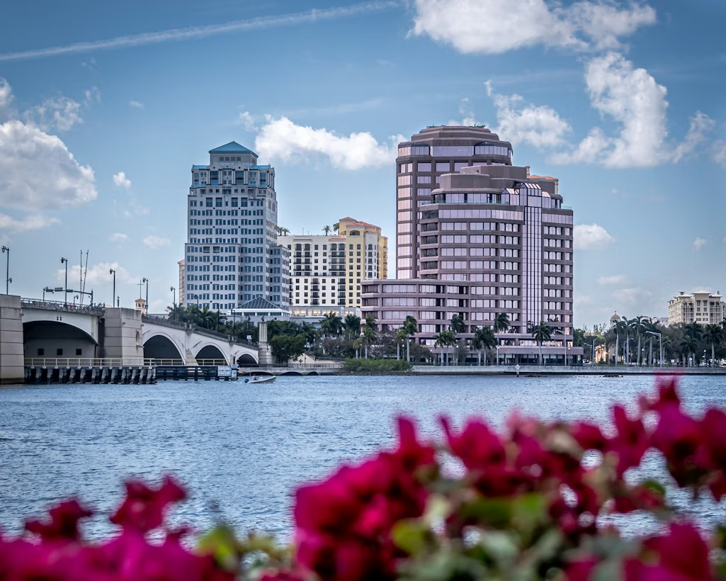 Waterfront view of West Palm Beach skyline with bridge and flowers in foreground.