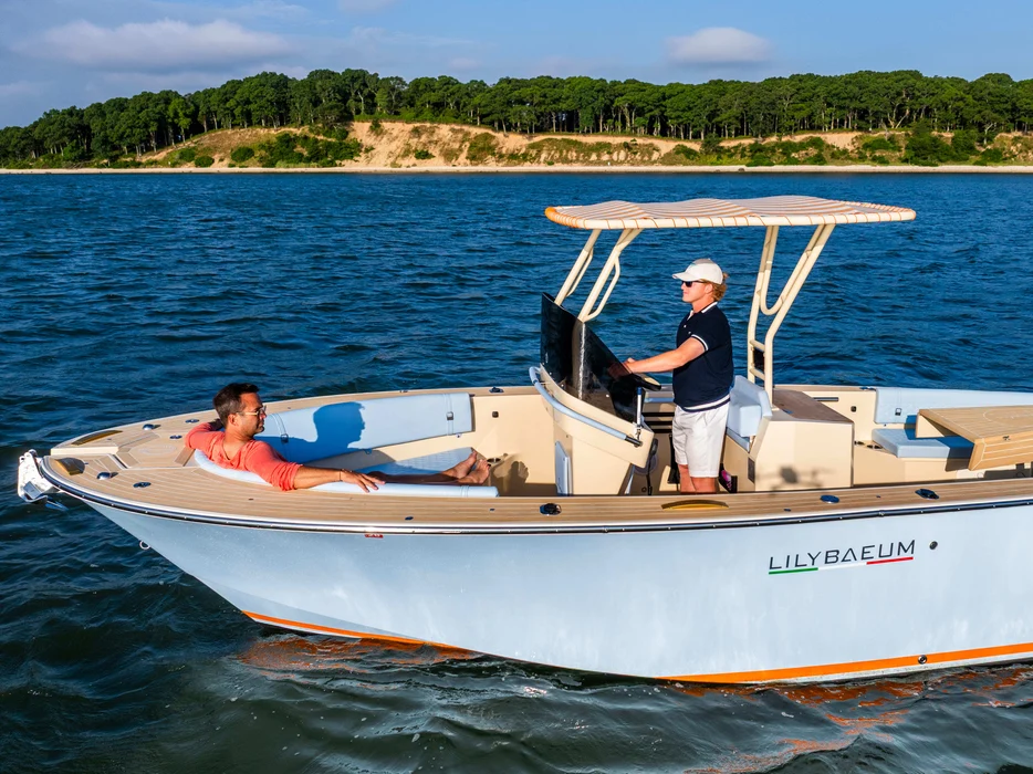 Two people cruising motorboat with forested shoreline backdrop.